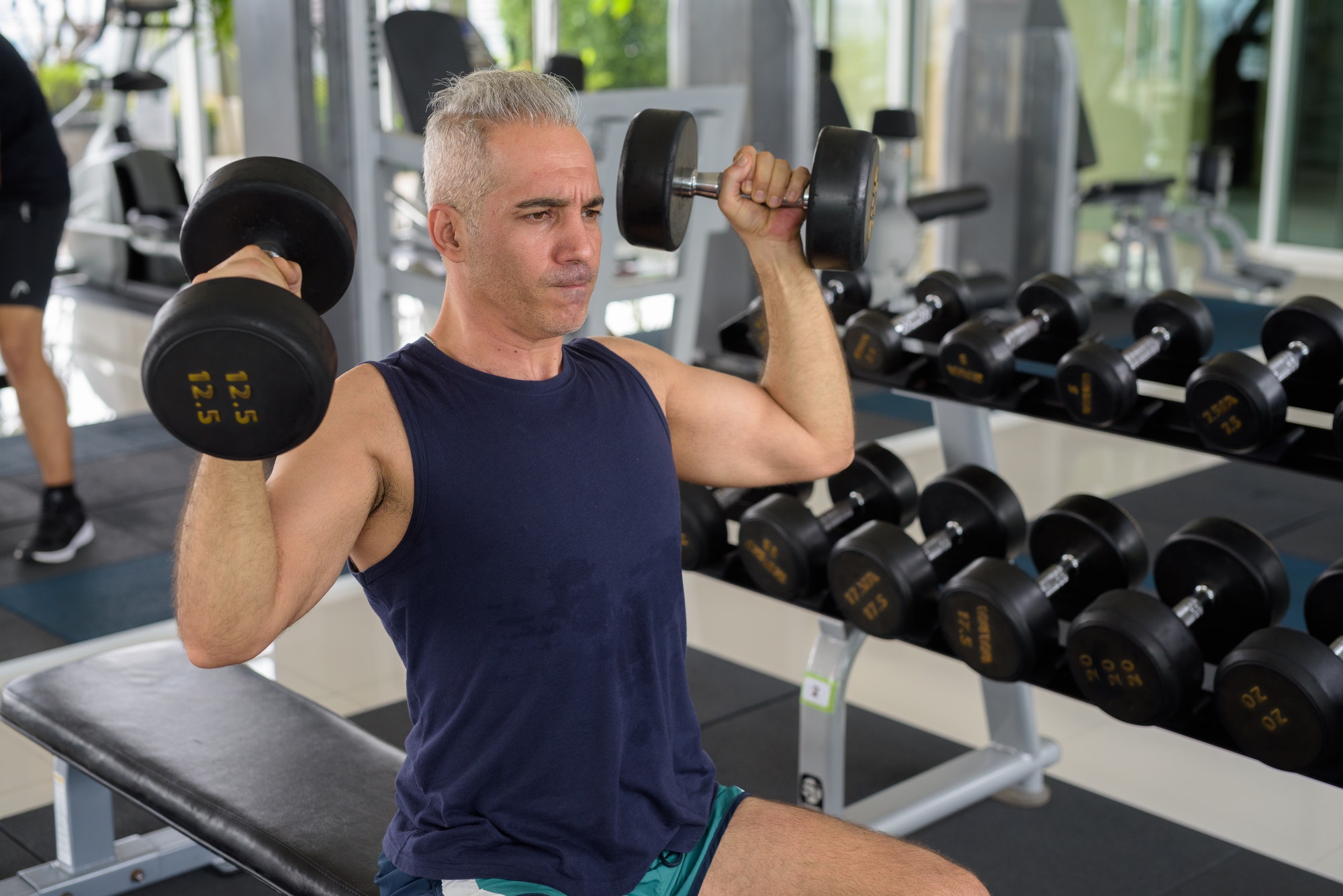 Mature handsome Persian man exercising with dumbbells at the gym